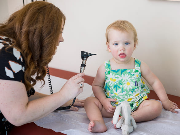 A baby sits on an exam table with a doctor going in for a hearing exam
