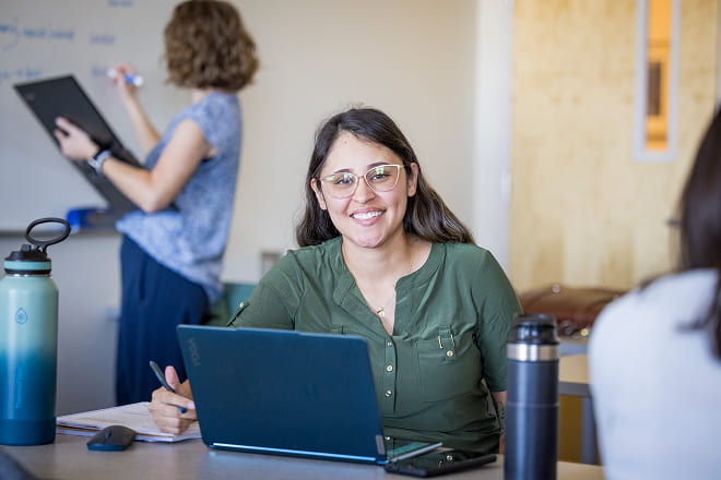 A students smiles while working on her laptop with other students nearby