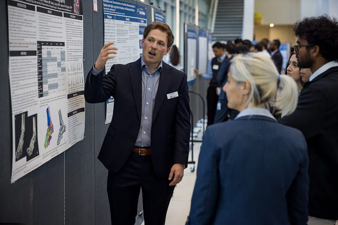 A student presents his research poster a group of onlookers
