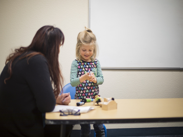 A young girl at a table with her teacher, engaged in a learning activity together.