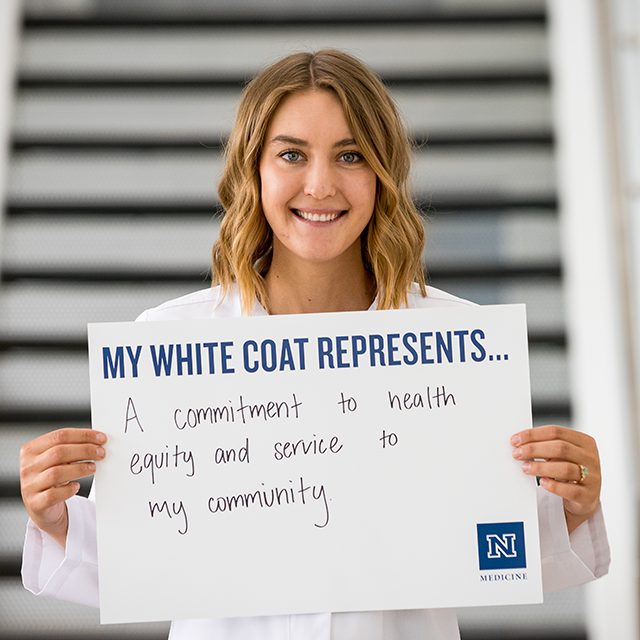 Sara Hanneman holding a sign that reads, "My White Coat represents... A commitment to health equity and service in my community."