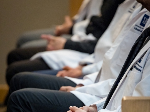 A lineup of medical students sitting together, dressed in white coats, appearing focused and attentive.