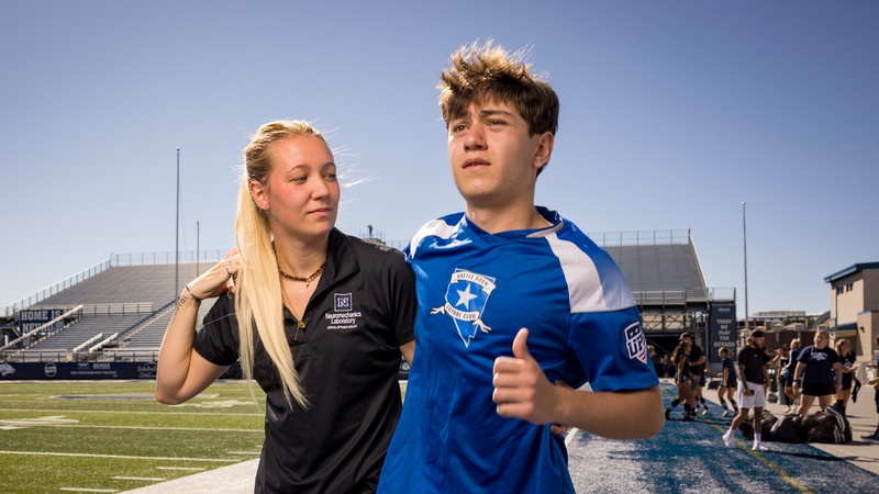 A medical student helps an athlete off a sports field while the athlete gives a thumbs up