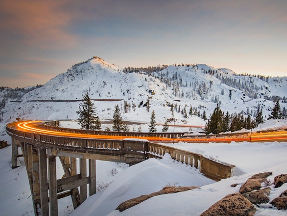 A snowy mountain bridge with a trail of lights from cars driving across it, surrounded by a winter landscape
