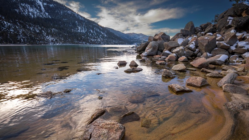 A serene lake with clear water and scattered rocks in the foreground, with snowy mountains in the background