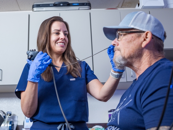 A female pathologist in blue scrubs and gloves uses a medical device to examine an older man's throat in a clinic, both smiling.