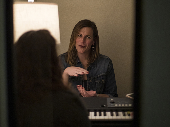 A pathologist in a denim jacket gesture expressively while talking, seated by a keyboard. A warm lamp glows beside her, creating a cozy atmosphere.