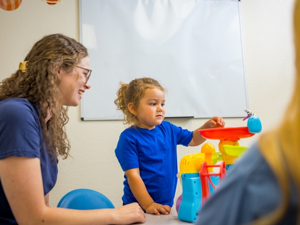 A pathologist and a young child playing with colorful toys at a table. The child looks focused, while the pathologist smiles supportively.