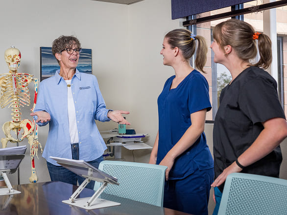 A professor standing in front of a skeleton model talks to two students