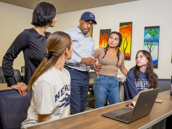 A diverse group of Speech-language pathology students and faculty having a casual discussion in a classroom. The setting is positive and engaged, with art on the walls and a laptop on the table.
