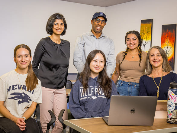 A group of professors and students around a laptop smile at the camera