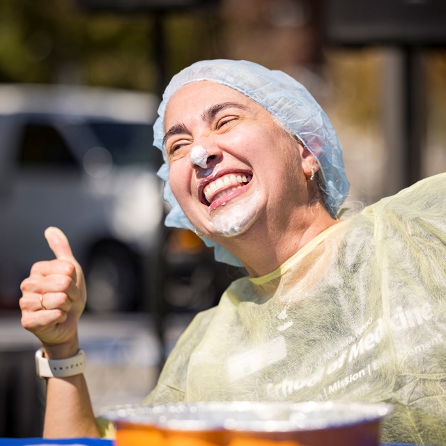 A faculty member smiles playfully at the camera with whipped cream on their face while giving a thumbs up
